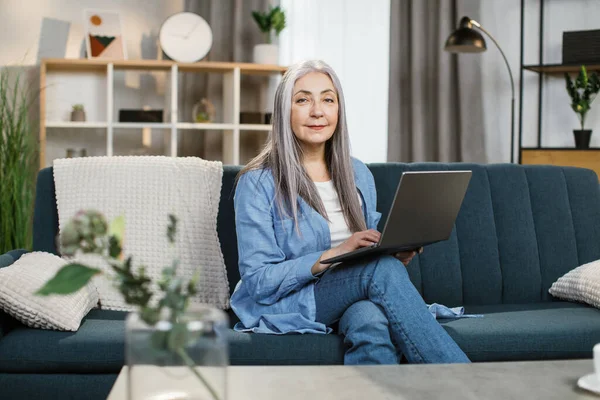 Pleasant attractive grey-haired woman with long hair sitting on couch with laptop on knees. — Stock Photo, Image