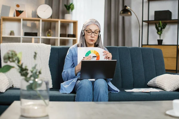 Attractive pleasant mature female sitting on sofa and showing graphs and charts. — Stock Photo, Image