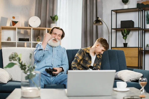 Anciano y su nieto sentados en el sofá y usando joysticks inalámbricos para jugar videojuegos —  Fotos de Stock