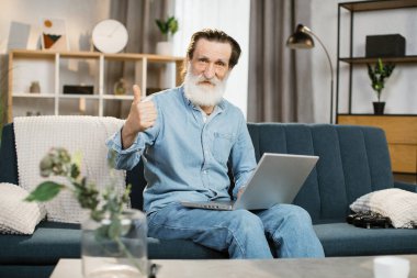 Evde oturma odasındaki kanepede oturup bilgisayarını video konuşması için kullanan yakışıklı bir beyefendi.