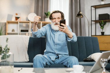 Gri sakallı, zarif adam rahat oturma odasındaki kanepede mutlu bir şekilde oturuyor.