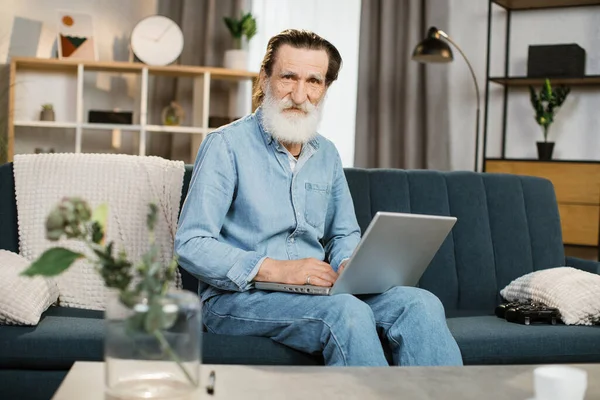 Evde oturma odasındaki kanepede oturup bilgisayarını video konuşması için kullanan yakışıklı bir beyefendi.