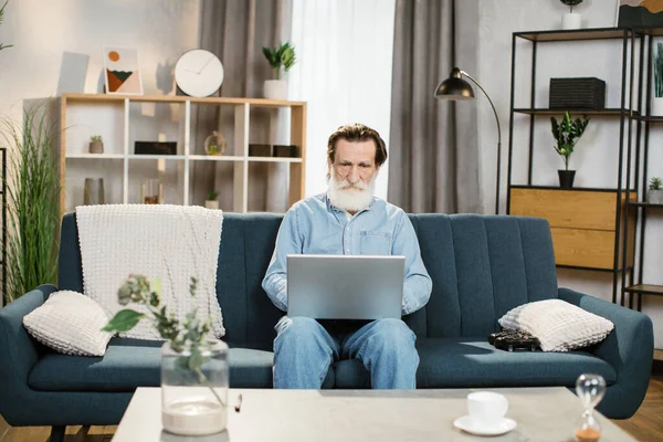 Άνδρας με γκρίζα γενειάδα ώριμος άνδρας σε casual ρούχα, κάθεται σε μπλε μαλακό καναπέ με laptop — Φωτογραφία Αρχείου