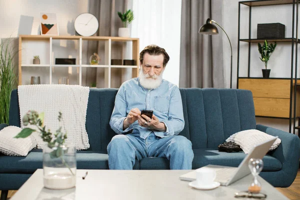 Senior γενειοφόρος άνδρας, κάθεται στον καναπέ στο άνετο σαλόνι σε εσωτερικούς χώρους και χρησιμοποιώντας smartphone — Φωτογραφία Αρχείου