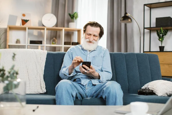 Hombre barbudo mayor, sentado en el sofá en la acogedora sala de estar en el interior y el uso de teléfonos inteligentes —  Fotos de Stock