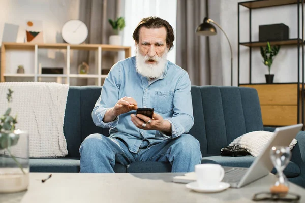 Senior άνθρωπος με έκπληξη έκφραση στο πρόσωπό του κάθεται στον καναπέ στο σπίτι έχοντας βιντεοκλήση — Φωτογραφία Αρχείου