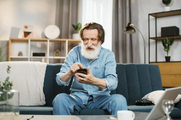 Hombre elegante confiado con barba gris sonriendo felizmente sentado en el sofá en la acogedora sala de estar —  Fotos de Stock