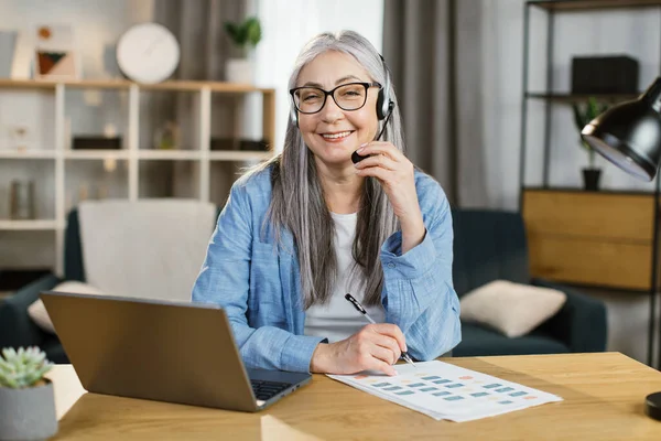 Femme plus âgée positive utilisant casque et ordinateur portable pour la conversation vidéo au bureau. — Photo