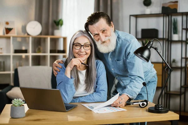 Θετική ηλικιωμένη σύζυγος και σύζυγος σε casual ένδυμα ποζάρουν στο φωτεινό σπίτι με σύγχρονα gadgets στο τραπέζι. — Φωτογραφία Αρχείου