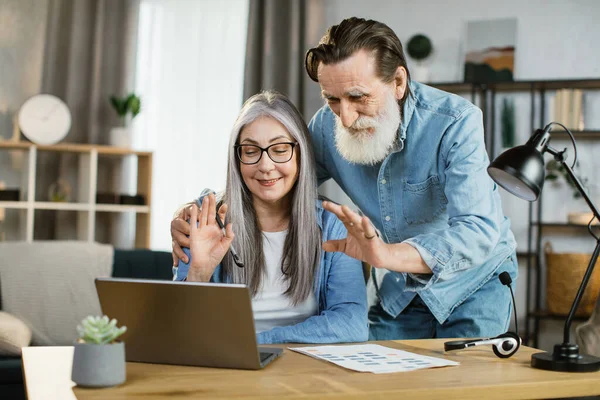 Positive senior wife and husband in casual wear posing at bright home with modern gadgets on table. — Stock Photo, Image