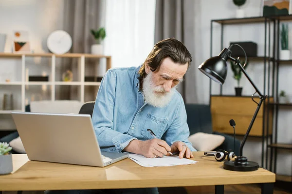 Ανώτερος γενειοφόρος άντρας κρατάει σημειώσεις ενώ πληκτρολογεί στο λάπτοπ στο γραφείο. — Φωτογραφία Αρχείου