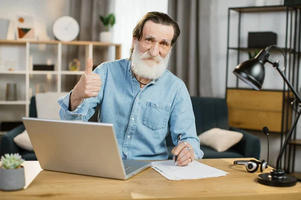 Idősebb férfi alkalmi viselet ül asztalnál modern laptop, jegyzetel és mosolyog a kamera. — Stock Fotó