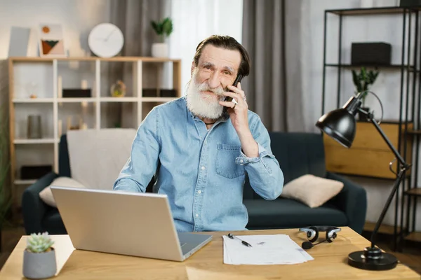 Ευχάριστη ώριμη Καυκάσιος επιχειρηματίας μιλάει στο κινητό και εργάζεται σε φορητό υπολογιστή στο τραπέζι. — Φωτογραφία Αρχείου