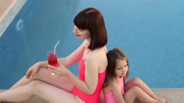 Genç anne ve küçük kızı aynı pembe mayolarıyla havuzun kenarında oturuyorlar..
