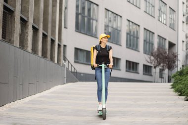 Sırt çantalı ve şapkalı kadın teslimatçı şehir yolu boyunca binalara karşı elektrikli scooter kullanıyor.