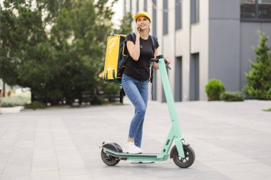 Şehirde elektrikli scooterla dururken akıllı telefon kullanan teslimatçı kadın..