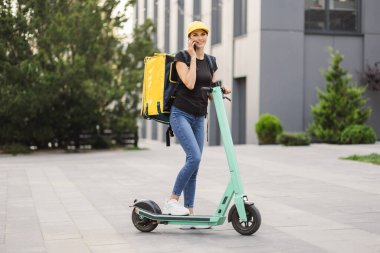 Şehirde elektrikli scooterla dururken akıllı telefon kullanan teslimatçı kadın..