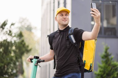 Sarı termal çantalı kurye yemek teslimatı elektrikli scooterla caddede yürüyor.