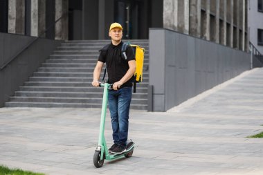 Sarı şapkalı ve termal yalıtkan çantalı mutlu teslimatçı. Şehir caddesinde elektrikli scooter kullanıyor..