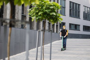 Sarı maskeli bir adam, şehirde sıcak yemek getiren bir scooter kullanıyor.