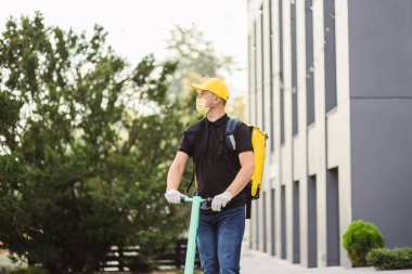Beyaz tenli teslimatçı, yüz maskeli elektrikli scooter 'la sarı termal çantalı..