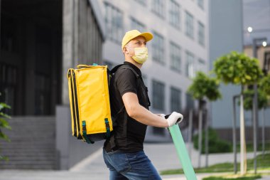 Beyaz tenli teslimatçı, yüz maskeli elektrikli scooter 'la sarı termal çantalı..