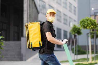 Beyaz tenli teslimatçı, yüz maskeli elektrikli scooter 'la sarı termal çantalı..