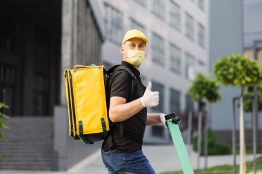 Beyaz tenli teslimatçı, yüz maskeli elektrikli scooter 'la sarı termal çantalı..