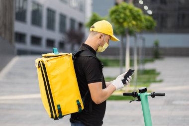 Akıllı telefon kullanan ve şehirde scooter kullanan yüz maskeli bir teslimatçının yan görüntüsü.
