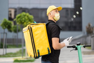 Akıllı telefon kullanan ve şehirde scooter kullanan yüz maskeli bir teslimatçının yan görüntüsü.