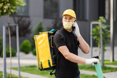 Sarı üniformalı teslimatçı elinde akıllı telefonuyla bir apartmanın arka planında duruyor.