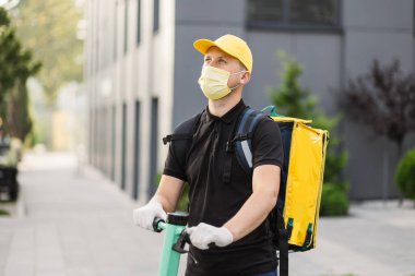 Beyaz tenli teslimatçı, yüz maskeli elektrikli scooter 'la sarı termal çantalı..