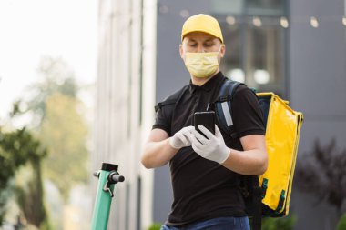 Akıllı telefon kullanan ve şehirde scooter kullanan yüz maskeli bir teslimatçının ön görüntüsü..