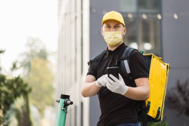 Akıllı telefon kullanan ve şehirde scooter kullanan yüz maskeli bir teslimatçının ön görüntüsü..