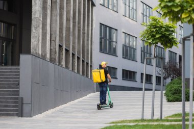 Sırtında sarı bir sırt çantası ve günlük yaz kıyafetleri olan genç bir adam elektrikli scooterla hareket ediyor.