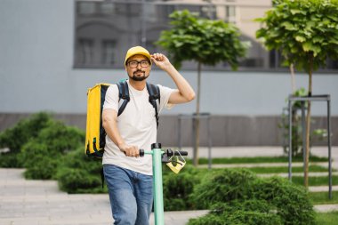 Beyaz erkek kurye scooterla şehirde sıcak yiyeceklerle yemek dağıtıyor.