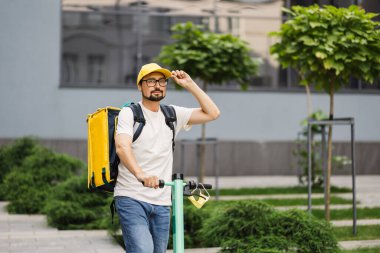 Beyaz erkek kurye scooterla şehirde sıcak yiyeceklerle yemek dağıtıyor.