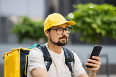 Sırtında büyük bir termal sırt çantasıyla akıllı telefondan konuşan beyaz bir yemek kuryesi.