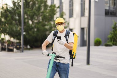 Sarı üniformalı teslimatçı elinde akıllı telefonuyla bir apartmanın arka planında duruyor.