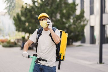 Sarı üniformalı teslimatçı elinde akıllı telefonuyla bir apartmanın arka planında duruyor.