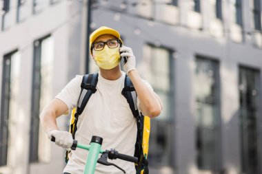 Sarı üniformalı teslimatçı elinde akıllı telefonuyla bir apartmanın arka planında duruyor.