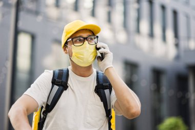 Sarı üniformalı teslimatçı elinde akıllı telefonuyla bir apartmanın arka planında duruyor.