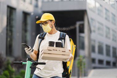 Güvenlik maskesi, eldiven ve üniformalı bir kurye elinde pizza kutuları ve kahve fincanları tutuyor.