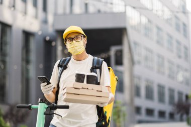 Koruyucu maskeli teslimatçı elinde bir yığın pizza karton kutusuyla iki fincan kahve tutuyor.