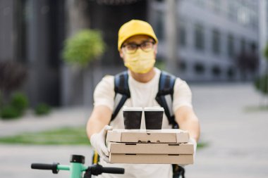 Koruyucu maskeli teslimatçı elinde bir yığın pizza karton kutusuyla iki fincan kahve tutuyor.