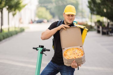 Sarı şapkalı ve termal yalıtımlı çantalı mutlu teslimatçı karton kutuda pizza ikram ediyor.