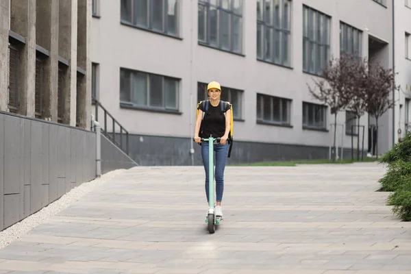 Lady delivery worker with backpack and cap rides electric scooter along city road against buildings — Stock Photo, Image