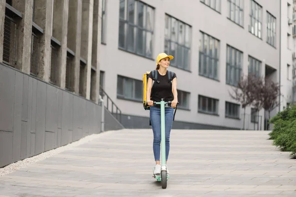 Sırt çantalı ve şapkalı kadın teslimatçı şehir yolu boyunca binalara karşı elektrikli scooter kullanıyor.