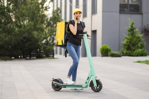 Şehirde elektrikli scooterla dururken akıllı telefon kullanan teslimatçı kadın..