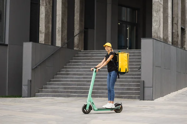 Tiro de comprimento total da jovem mulher de entrega em boné amarelo com saco térmico ou mochila montando uma scooter — Fotografia de Stock